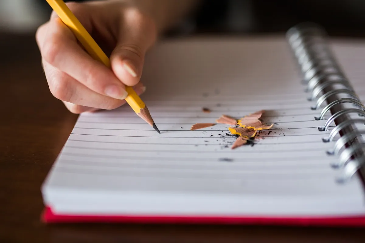 Mão escrevendo com lápis em caderno espiral durante estudo de notas e médias