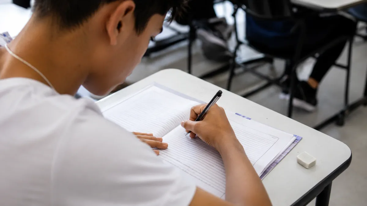 Estudante escrevendo em folha em mesa de sala de aula representando redação do ENEM